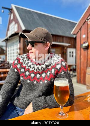 Porträt eines jungen Mannes in einem isländischen Wollpullover, der im Juli ein Bier auf der Terrasse des Restaurants in Husavik genießt. Stockfoto