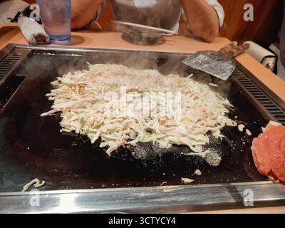 Traditionelles japanisches Okonomiyaki, das auf einem Teppan-Grill zubereitet wird, mit köstlicher Textur und authentischem Street Food-Stil. Stockfoto