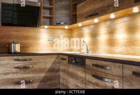 Moderne Eckküche aus hellbraunem Holz mit passendem Backsplash, eingebautem Waschbecken, Dunstabzugshaube und Topf auf dem Herd, warm beleuchtet durch die Unterkabine Stockfoto