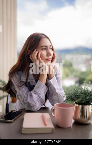 Eine ruhige asiatische Erwachsene Frau auf einem Balkon genießt einen friedlichen Moment und blickt nachdenklich auf die Panoramastadt und Berglandschaft Stockfoto
