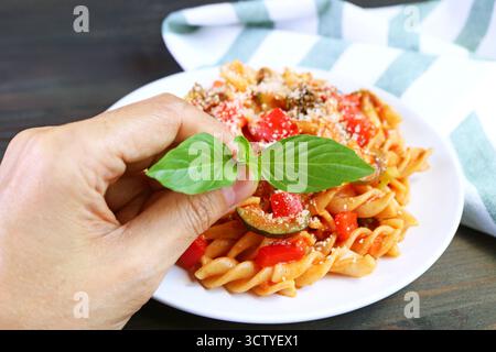 Geben Sie Basilikumblätter von Hand auf ein Gericht mit frisch gekochtem Vollkorn-Fusilli-Pasta in Tomatensauce Stockfoto