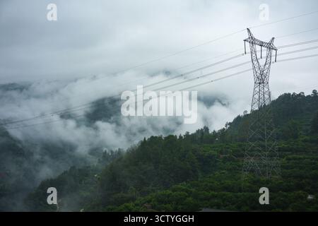 Ein hoher Metallturm steht auf einem grünen, bewaldeten Berg und trägt Stromleitungen über ein nebeliges Tal. Stockfoto