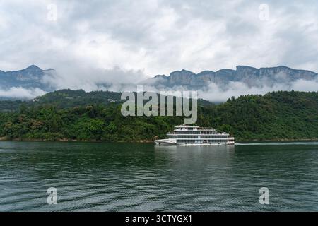 Ein großes weißes Boot segelt auf einem ruhigen Fluss mit grünen nebeligen Bergen im Hintergrund in Yichang City China. Stockfoto