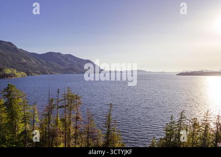 Ruhiges Wasser reflektiert den Himmel, während die Sonne hinter fernen Bergen untergeht. Hohe Bäume säumen das Ufer und schaffen eine ruhige Atmosphäre in der Landschaft. Stockfoto