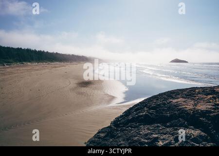 Sanfte Wellen tummeln sich am Sandstrand, während Surfer auf dem Wasser reiten. Ein felsiger Felsvorsprung steht im Kontrast zum glatten Strand und den Bäumen in der Ferne an einem sonnigen Tag. Stockfoto
