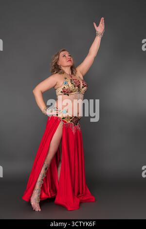 Ernsthafte Erwachsene Tänzerin mittleren Alters mit handgezogenem Anzug im Tanzkostüm - BH-Oberteil und scharlachroter langer Rock mit Schlitz bewegt sich barfuß in voller Länge Stockfoto