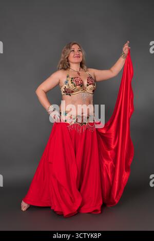 Lächelnde und glückliche kurvige Tänzerin, die in Tanzkostüm kleidet - BH-Oberteil und roter Maxirock, sie trägt keine Schuhe und tritt in voller Länge auf Stockfoto