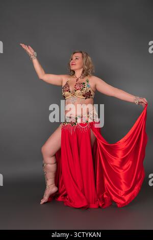 Lächelnde Tänzerin in voller Länge vor grauem Hintergrund. Erwachsene Frau mittleren Alters kleidet sich in Tanzkostüm - BH-Top, roter langer Rock mit Stockfoto