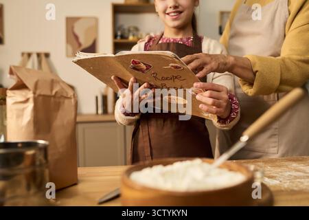 Kaukasisches Mädchen und Frau mittleren Alters bereiten zusammen in der Küche Essen zu, halten Rezeptbuch und befolgen Anweisungen, beide tragen Schürzen, Mehl und Backzutaten auf dem Tisch Stockfoto