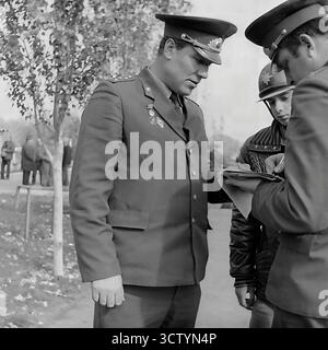 Ein ehrliches Porträt eines sowjetischen GAI-Beamten bei der Arbeit auf diesem authentischen Schwarzweiß-Foto aus den 1970er Jahren Der uniformierte Miliz, der Dienstausweise auf der Brust trägt, konzentriert sich sorgfältig auf das Ausfüllen von Papieren, wahrscheinlich während einer Fahrzeuginspektion bei einem MREO in Sloviansk, Ukrainische SSR. Das Bild bietet eine Nahaufnahme einer Autorität im dokumentarischen Stil, die die bürokratische Realität und das ernste Verhalten der Strafverfolgungsbehörden im täglichen Leben der Sowjetunion erfasst Stockfoto