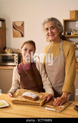Kaukasische Seniorin und kaukasisches Mädchen stehen zusammen in der Küche und lächeln in die Kamera, halten frisch gebackenes Brot auf einem Tablett, beide tragen Schürzen und zeigen die Backtätigkeit der Familie Stockfoto