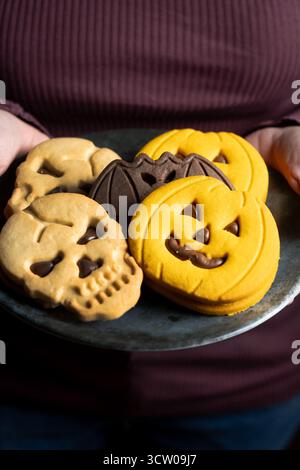 Hände halten Teller mit Halloween-Keksen in Form von Kürbissen, Schädeln und Fledermäusen auf Holztisch Stockfoto