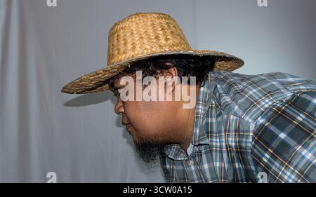 Ein indonesischer Mann trägt einen Strohhut und ein grünes kariertes Hemd auf weißem Hintergrund. Stockfoto