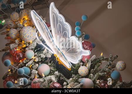 Die Nahansicht zeigt einen wunderschön verzierten Weihnachtsbaum mit einem auffälligen, beleuchteten Schmetterlingsschmuck, der sanft leuchtet und eine Uni hinzufügt Stockfoto