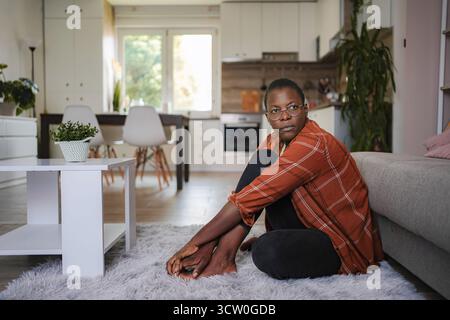 Junge Frau, die auf dem Boden sitzt und nachdenklich in ihrer modernen Wohnung aussieht Stockfoto