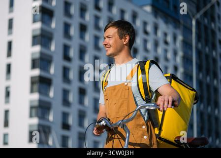 Ein glücklicher Kurier, der in der Stadt arbeitet, eine hellgelbe Liefertasche trägt und ein Fahrrad schiebt. Er verkörpert wichtige Themen wie Stadtservice, Nachhaltigkeit und Gig Economy Stockfoto