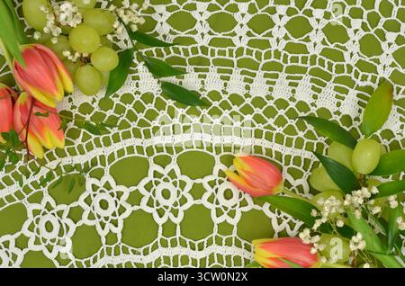 Große, weiße, Vintage, gehäkelte Tischdecke auf grünem Hintergrund, verziert mit Tulpen und Trauben. Stockfoto