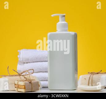 Weiße quadratische Flasche mit Spender und Stapel gefalteter weißer Handtücher, satter gelber Hintergrund. Körperpflege oder Spa-Behandlungskonzept Stockfoto