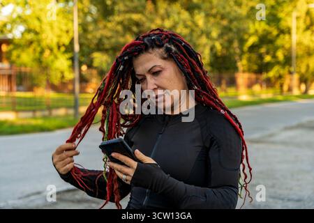 Frau mit roten Dreadlocks mit Smartphone in einem Park, in Sportkleidung Stockfoto