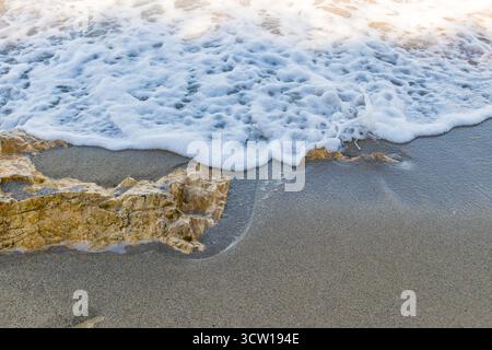 Meereswelle, die bei Sonnenuntergang über gelben Felsen am Sandstrand weht. Nahaufnahme der Surftextur des Ozeans und der Natur an der Küste. Stockfoto
