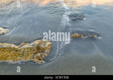 Meereswelle, die bei Sonnenuntergang über gelben Felsen am Sandstrand weht. Nahaufnahme der Surftextur des Ozeans und der Natur an der Küste. Stockfoto