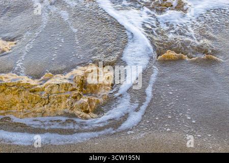 Meereswelle, die bei Sonnenuntergang über gelben Felsen am Sandstrand weht. Nahaufnahme der Surftextur des Ozeans und der Natur an der Küste. Stockfoto