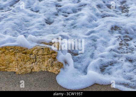 Meereswelle, die bei Sonnenuntergang über gelben Felsen am Sandstrand weht. Nahaufnahme der Surftextur des Ozeans und der Natur an der Küste. Stockfoto