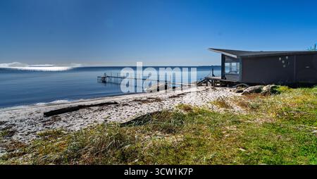 Eine ruhige Küstenszene mit einem modernen Strandhaus, einem hölzernen Dock, der sich in ruhiges Wasser erstreckt, und einer Sandküste mit grasbewachsenen Dünen unter einem klaren Wasser Stockfoto