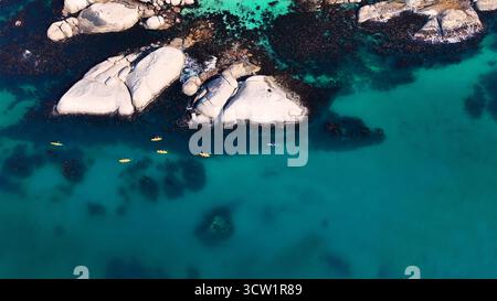 Aus der Vogelperspektive auf den türkisfarbenen Ozean, der um Granitfelsen wirbelt, während Kajaks entlang der Küste in der Nähe von Llandudno Beach, Kapstadt, Westkap, Südafrika treiben. Stockfoto