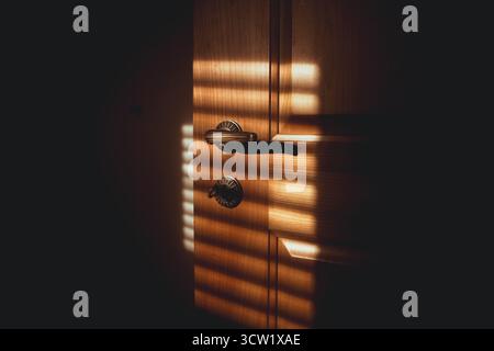 Sonnenlicht auf einer hölzernen Wohnzimmertür und Messinggriff. Nahaufnahme, Weitwinkelaufnahme, dunkler, stimmungsvoller Hintergrund, keine Leute Stockfoto