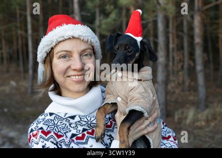 Fröhliche Frau in einem Weihnachtspullover hält einen kleinen schwarzen Hund, während sie ihn im Wald spaziert. Eine Frau und ihr Haustier in roten Santa-Hüten. Wir feiern das neue Y Stockfoto