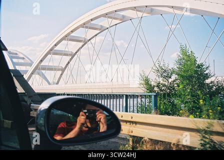 Analoges 35-mm-Foto vom Beifahrersitz mit der Žeželj-Brücke in Novi Sad, mit einem Fotografen im Rückspiegel, der eine Filmkamera hält. Stockfoto
