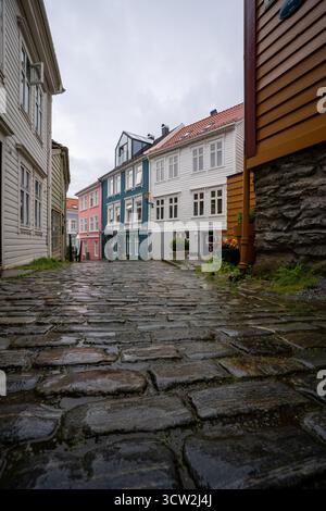 Flacher Blick auf eine kopfsteingepflasterte Straße im historischen Zentrum von Bergen, Norwegen, mit farbenfrohen Holzhäusern und einem Touristen mit einem Sonnenschirm bei einem Regen Stockfoto