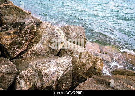 Große Felsbrocken am Ufer, die von den Wellen des türkisfarbenen Meeres oder Sees gewaschen werden, bilden eine raue Küstenlandschaft. Stockfoto