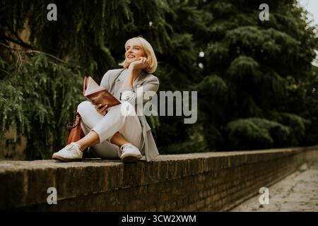 Eine Frau sitzt auf einer Steinmauer in einem Park und lächelt, während sie ein Buch liest. Sie trägt ein leichtes Outfit und genießt die friedliche Umgebung mit Bäumen A Stockfoto
