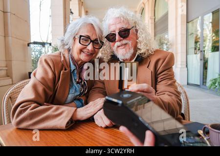 Ein glückliches Seniorenpaar lächelt weit, wenn es mit seiner Kreditkarte bezahlt, und hebt seine Gesellschaft und Freude hervor Stockfoto