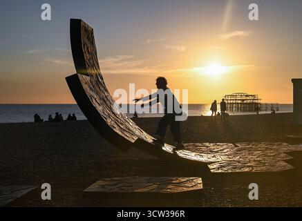Brighton UK 9 Oktober 2025 - Besucher genießen einen wunderschönen Sonnenuntergang über Brighton Beach Passacaglia Skulptur und West Pier nach einem weiteren warmen Tag entlang der Südküste . Passacaglia ist eine riesige, gekrümmte abstrakte Skulptur von Charles Hadcock, die aus recyceltem Gusseisen gebaut wurde, 20 Tonnen schwer und 5 Meter hoch ist: Credit Simon Dack / Alamy Live News Stockfoto