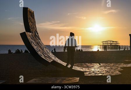 Brighton UK 9 Oktober 2025 - Besucher genießen einen wunderschönen Sonnenuntergang über Brighton Beach Passacaglia Skulptur und West Pier nach einem weiteren warmen Tag entlang der Südküste . Passacaglia ist eine riesige, gekrümmte abstrakte Skulptur von Charles Hadcock, die aus recyceltem Gusseisen gebaut wurde, 20 Tonnen schwer und 5 Meter hoch ist: Credit Simon Dack / Alamy Live News Stockfoto