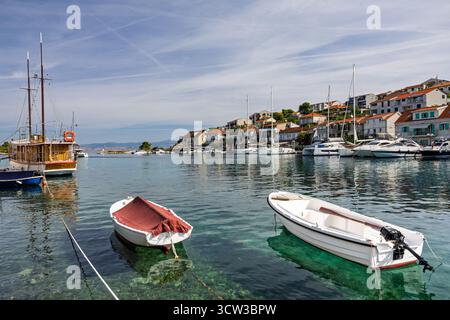 Eine ruhige Hafenszene mit kleinen Ruderbooten, vertäuten Segelbooten und einem Dorf am Meer mit roten Ziegeldächern. Ruhiges Wasser reflektiert die Gefäße und Stockfoto