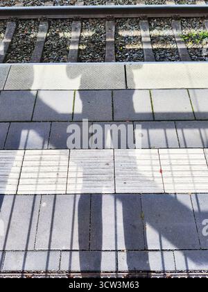 Langer Schatten einer Person, die auf einem texturierten Bahnsteig in der Nähe von Eisenbahngleisen steht Stockfoto