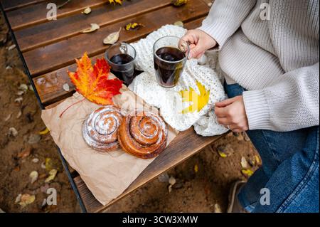 Nahaufnahme von Frauenhänden in Strickpullover, die eine gemütliche Herbstkaffeepause mit Gebäck und bunten Herbstblättern im Freien genießen Stockfoto