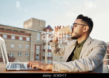 Junger attraktiver Mann aus dem Nahen Osten, der Gläser trägt und Kaffee aus dem Pappbecher weiß trinkt, sitzt am Tisch mit Laptop auf der Dachterrasse des Cafés Stockfoto