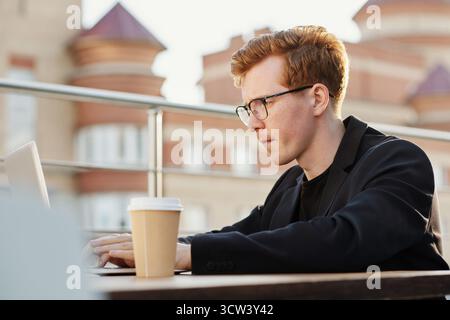 Junger konzentrierter rothaariger kaukasischer Mann, der am Laptop arbeitet, während er am Kaffeetisch auf der Dachterrasse auf einem unscharfen Hintergrund sitzt Stockfoto