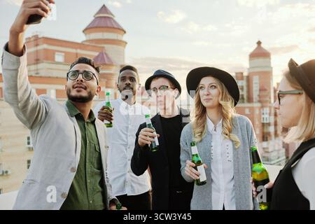 Junger Mann aus dem Nahen Osten, der Selfie auf dem Smartphone macht, mit seinen trendigen männlichen und weiblichen Freunden, die mit Bier auf der Dachterrasse rumhängen Stockfoto
