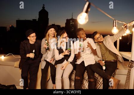 Multiethnische Gruppe junger, stylischer männlicher und weiblicher Freunde, die Selfie auf dem Smartphone machen, während sie am Sommerabend mit Bier auf dem Dach abhängen Stockfoto