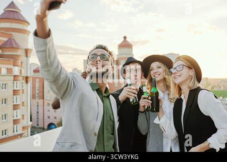 Multiethnische Gruppe junger, stilvoller männlicher und weiblicher Freunde, die Selfie auf dem Smartphone machen, während sie auf der Dachterrasse mit Bier abhängen Stockfoto