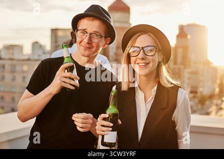 Terrasse bei Sonnenuntergang. Porträt eines jungen, stylischen, hippigen kaukasischen Mannes und einer Frau, die auf dem Dach stehen, Bier trinken und in die Kamera lächeln Stockfoto