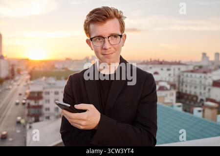 Porträt eines jungen, attraktiven rothaarigen Kaukasiers, der mit Smartphone auf dem Dach steht und nachdenklich wegschaut Stockfoto