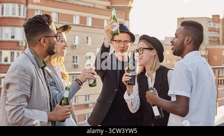 Multiethnische Gruppe junger, trendiger männlicher und weiblicher Kollegen, die Bier trinken, sich unterhalten und den Abend auf der Dachterrasse genießen Stockfoto