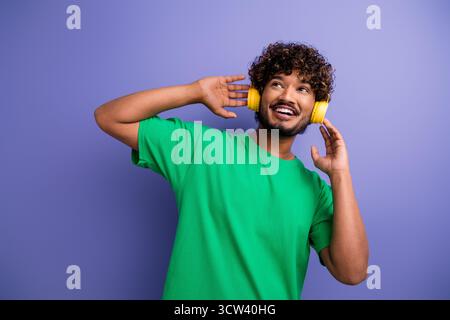 Positiver Mann in einem grünen T-Shirt, der Musik mit gelben Kopfhörern genießt und aufgeregt vor einem leuchtend violetten Hintergrund posiert Stockfoto